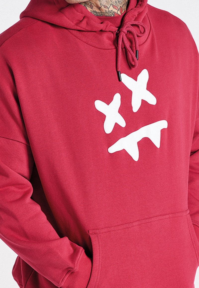 Sweat à capuche rouge en tissu doux arborant un grand motif blanc avec deux X et un motif en zigzag. Comprend une poche avant et une capuche.