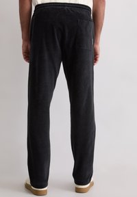 Mann trägt schwarze Cordhosen mit elastischem Bund und Gesäßtasche, kombiniert mit weißen Sneakers und einem in die Hose gesteckten weißen Hemd.