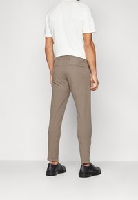 Man står vänd bort med slim-fit beige byxor, vit kortärmad skjorta och svarta läderskor med snörning mot en enkel bakgrund.