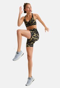 Top sportivo nero a maniche corte e pantaloni corti coordinati con motivi astratti gialli. Scarpe da corsa grigie con dettagli azzurri. Modella in movimento.
