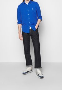 Niebieska koszula z długim rękawem z guzikami, ciemne dżinsy i białe buty sportowe z niebieskimi akcentami; luźny krój i podwinięte mankiety na dżinsach.