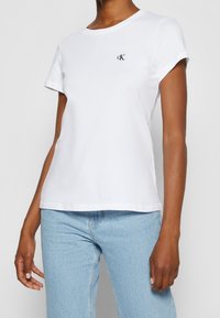 Hvid bomulds t-shirt med rund halsudskæring og korte ærmer, med et lille sort logo på venstre brystområde. Sammen med lyseblå jeans.
