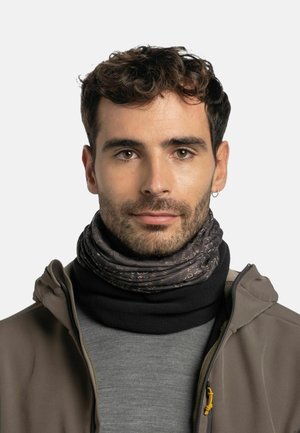 Schwarzer und gemusterter Schal, der unter einem grauen Hemd und einer braunen Jacke getragen wird. Der Schal weist ein geometrisches Muster in Brauntönen und Creme auf.