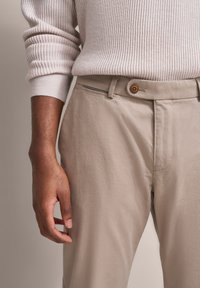 Eine dunkelhäutige Person trägt einen beigen, gerippten Pullover, der in beigen Hosen mit einem braunen Knopf und einer Reißverschlusstasche gesteckt ist.