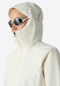 Giacca impermeabile di colore chiaro con cappuccio, zip completa e dettagli con logo. Abbinata a occhiali da sole grigi con un design sportivo.