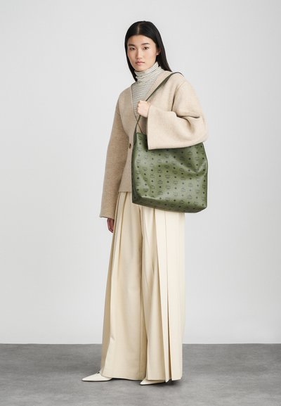 Olive green tote bag with a textured surface and patterned design, featuring a single shoulder strap. Model wears a beige sweater and cream pants.