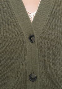 Olivově zelený pletený cardigan s výstřihem do V, vroubkovanou strukturou a dvěma černými knoflíky vpředu. Měkký materiál s viditelnými švy.