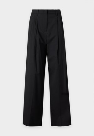 Femme aux longs cheveux marron portant un haut marron à manches courtes et un pantalon noir à jambes larges, debout avec une main dans la poche.