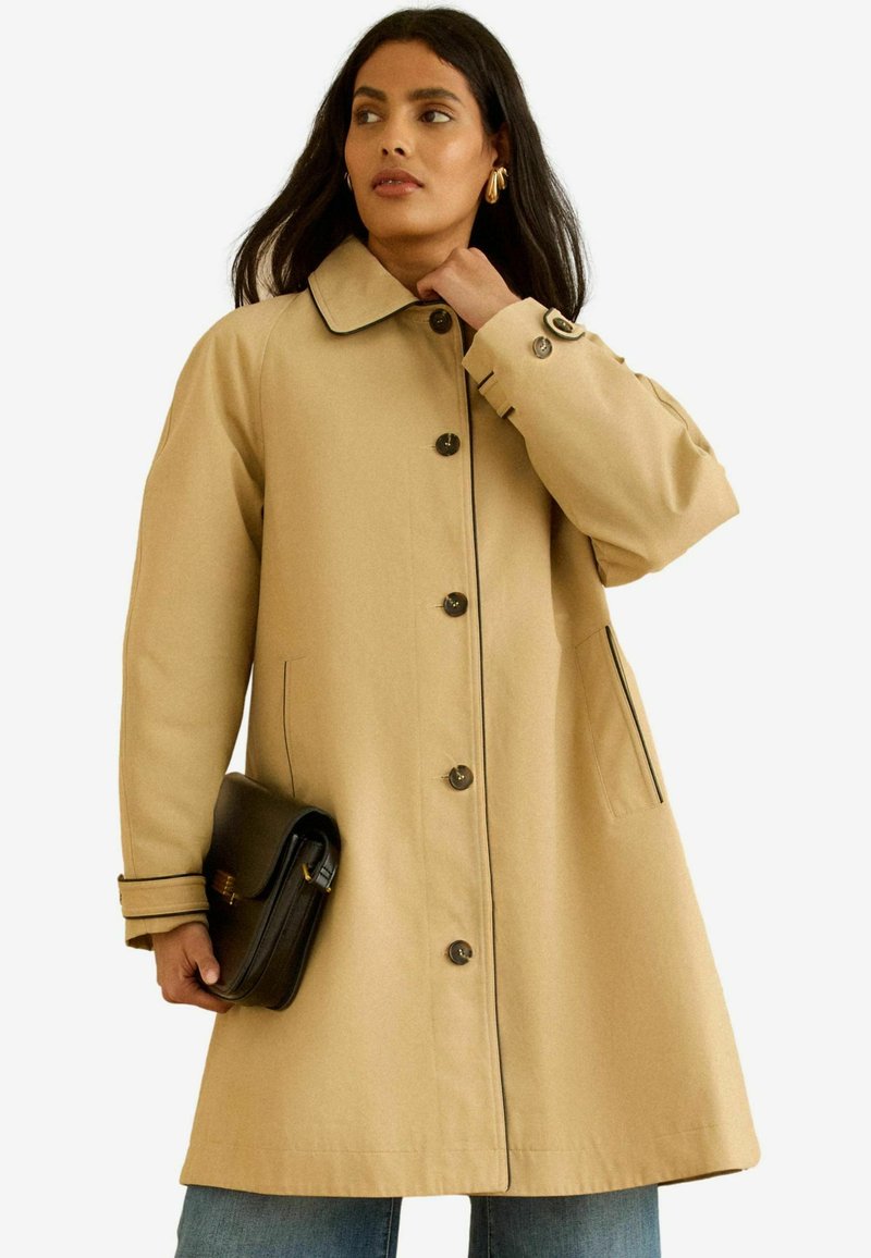 Femeie purtând o haină bej cu nasturi și bordură neagră, ținând o geantă plic neagră, privind în lateral, cu părul lung și întunecat și cercei aurii rotunzi.