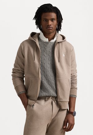 Felpa beige con zip e coulisse, indossata sopra un maglione grigio a treccia. Abbinata a joggers beige coordinati. Orologio al polso.