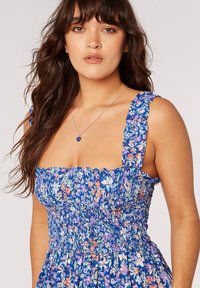 Blaues Blumenkleid mit gesmocktem Oberteil, Rüscheträgern und einem lebhaften Muster aus orangefarbenen und weißen Blumen auf einem strukturierten Stoff.