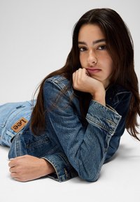 Mladá žena s dlhými tmavými vlasmi v modrej rifľovej bunde a džínsoch si opiera bradu o ruku a leží na svetlosivom povrchu.
