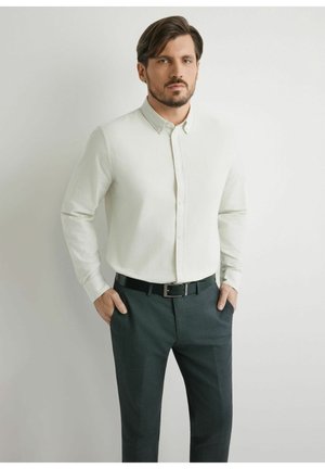 Mężczyzna o ciemnych włosach i brodzie, ubrany w białą koszulę na guziki oraz ciemnoszare spodnie, z rękami w kieszeniach, na jednolitym tle.