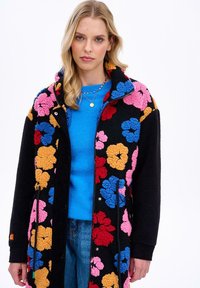 Čierny flísový kabát s viacfarebnými kvetinovými vzormi v ružovej, červenej, modrej a oranžovej farbe. Má zavinovací golier a zips.