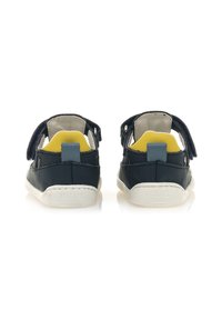 Sandalias de cuero azul marino con suelas blancas, parte trasera abierta y correas ajustables. Acentos amarillos y grises visibles en el área del talón.