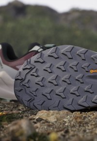 Nahaufnahme der Sohle eines Trailrunning-Schuhs mit tiefen, kantigen Stollen für Halt, die auf felsigem Gelände im Freien liegt.