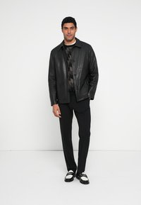 Schwarze Lederjacke mit Knopfverschluss, getragen über einem gemusterten Pullover, kombiniert mit maßgeschneiderten schwarzen Hosen und Schuhen mit weißen Akzenten.