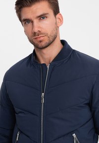 Bunda bomber v námořnické modři se zipem vpředu, rovnými manžetami a šikmým prošívaným vzorem. Vyrobena z hladké tkaniny a má dvě boční kapsy.