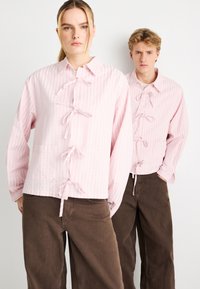 Ροζ ανοιχτόχρωμα ριγέ σακάκια με κουμπιά και δεσίματα, με άνετη εφαρμογή και μακριά μανίκια, συνδυασμένα με σκούρα καφέ φαρδιά παντελόνια.