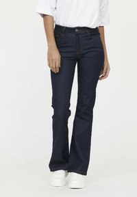 Donkerblauwe flared jeans van denim; heeft een hoge taille, een vijfzakensysteem en zichtbare oranje stiksels. Gestyled met witte sneakers.