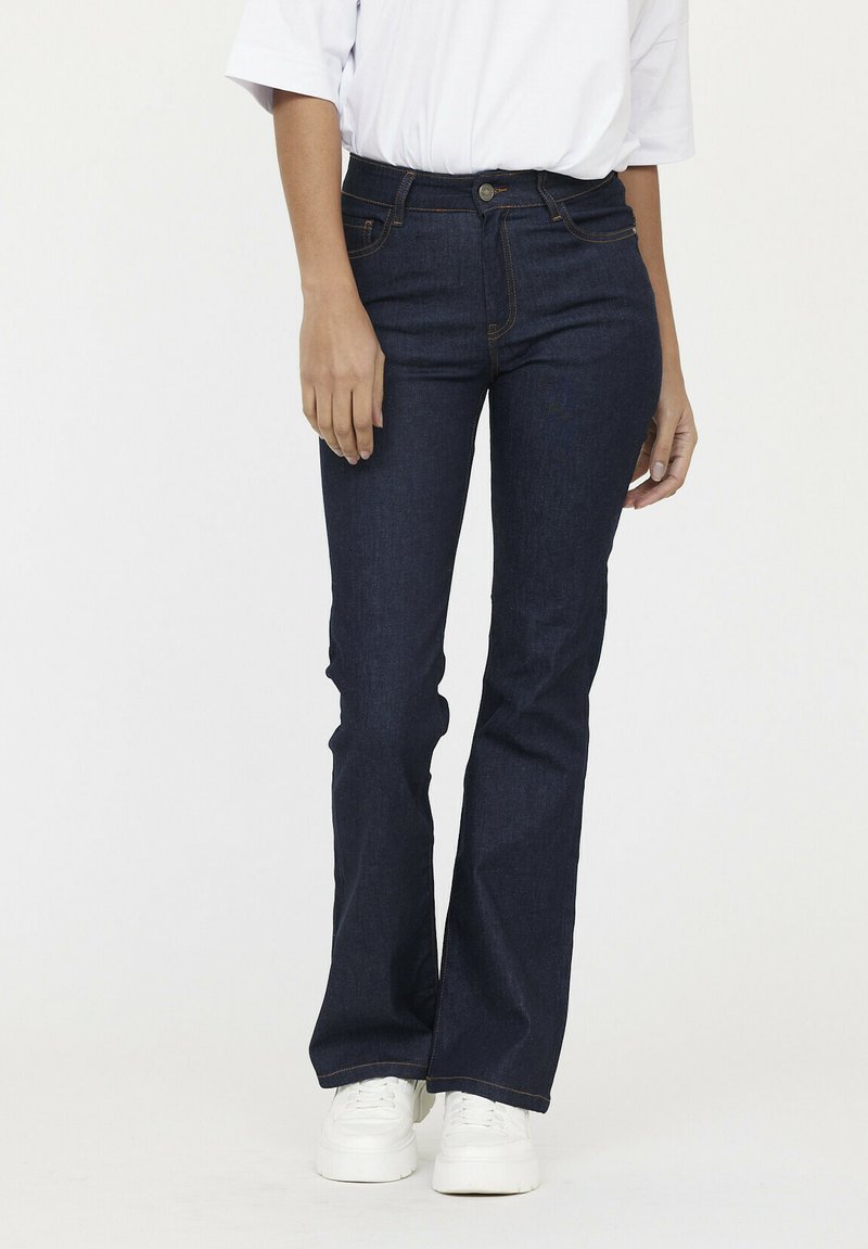 Donkerblauwe flared jeans van denim; heeft een hoge taille, een vijfzakensysteem en zichtbare oranje stiksels. Gestyled met witte sneakers.