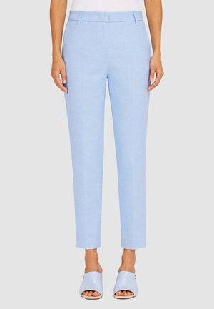 Donna che indossa pantaloni azzurri su misura a gamba dritta e scarpe azzurre aperte davanti, in piedi contro uno sfondo semplice.