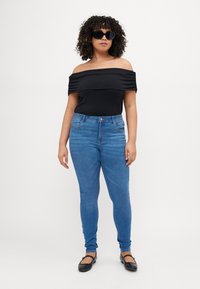 Sieviete ar cirtainiem matiem, ģērbusies melnā off-shoulder topā, zilās šaurās džinsās, melnās plakanās kurpēs un lielās melnās saulesbrillēs, stāv pretī baltai fona.