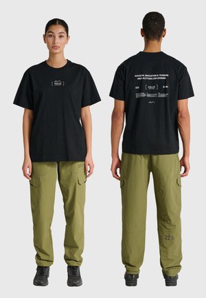 For- og bagfra billeder af to personer iført sorte grafiske t-shirts og olivengrønne cargobukser med sorte sneakers, stående mod en hvid baggrund.