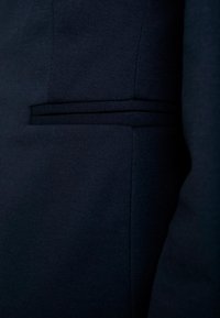 Námořnická svrchní bunda vyrobená z texturované látky, která má úzkou kapsu s klopou a čisté, upravené linie. Hladký povrch s minimálním zdobením.