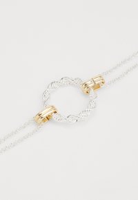 Pulseira de corrente prateada destacando um centro circular torcido, acentuada com componentes em tom de ouro liso. Acabamentos texturizados e polidos.