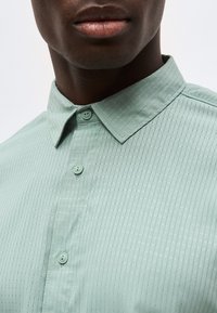 Κοντινό πλάνο ενός ατόμου που φοράει ένα ανοιχτοπράσινο πουκάμισο με κουμπιά, διακριτικό καρό σχέδιο και μυτερό γιακά.