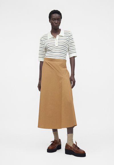 Γυναίκα που φοράει λευκό ριγέ πλεκτό πουκάμισο, μπεζ midi φούστα σε σχήμα Α, μπεζ κάλτσες και καφέ παπούτσια με κορδόνια και πάτο, στέκεται σε λευκό φόντο.