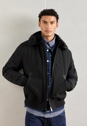 Chaqueta bomber negra con cuello de peluche, cierre delantero con cremallera y puños elásticos. Llevada sobre una camisa de mezclilla con un patrón de cuadros.