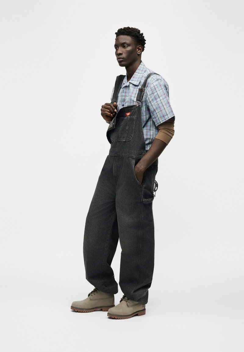 Giovane uomo che indossa una salopette di denim nera, una camicia a quadri con maniche lunghe marroni sotto, e stivali beige, in piedi con una mano in tasca.