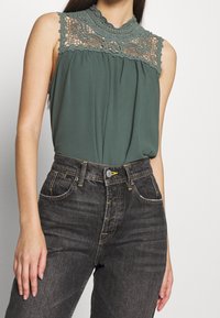 Blusa verde senza maniche con pizzo sul collo. Abbinata a jeans grigio scuro con cuciture gialle e chiusura con bottone.
