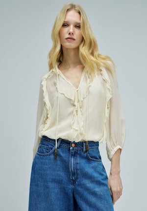 Mujer rubia con una blusa crema con volantes y lazos y vaqueros azules de talle alto sobre un fondo gris liso.
