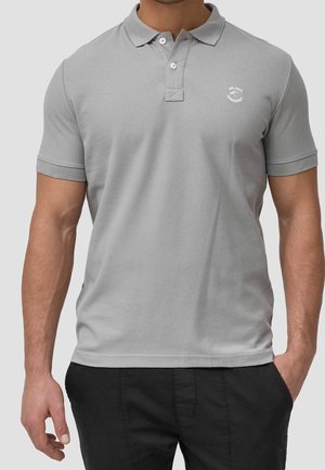 Homme portant un polo gris clair à manches courtes avec un col et deux boutons, associé à un pantalon noir, debout devant un fond uni.