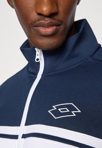 Blaue Zip-Jacke mit weißen Streifen und einem Logo, ausgestattet mit einem hohen Kragen und einer glatten Textur. Die Hardware umfasst einen weißen Reißverschluss.