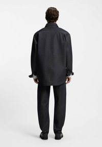 Uomo in piedi di spalle con giacca di jeans scura e pantaloni abbinati, indossa scarpe nere su sfondo bianco semplice.