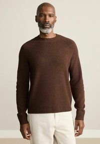 Brauner Strickpullover mit Rundhalsausschnitt, gerippten Bündchen und Saum. Der Stoff wirkt weich mit einer strukturierten Oberfläche und wird durch hellfarbige Hosen ergänzt.