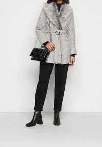 Abrigo de mezcla de lana gris con un cuello de piel sintética, cintura ceñida con un cinturón, combinado con pantalones negros y un bolso negro acolchado. Botines negros completan el look.