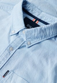 Jasnoniebieska koszula zapinana na guziki z wykładanym kołnierzykiem, z białymi guzikami, teksturowanym materiałem i małą metką marki na kieszeni.