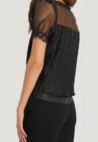 Schwarze Bluse mit durchsichtigen Puffärmel, plissierter Textur und einem soliden Unterband. Sie verfügt über einen runden Ausschnitt und dezente Rüschendetails an den Kanten.