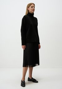 Schwarzer Strickpullover mit einem lockeren Schnitt, kombiniert mit einem schwarzen Faltenrock und schwarzen Slippern mit dekorativen Hardware-Details.