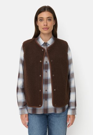 Kvinde iført en brun fleecervest over en blå og brun ternet skjorte med blå jeans, stående mod en ensfarvet hvid baggrund.