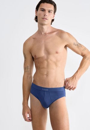 Shirtloze man met tatoeages die blauwe Sloggi slipjes aanpast tegen een effen lichte achtergrond.