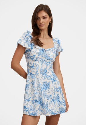 Frau mit langen braunen Haaren, die ein weißes kurzes Kleid mit blauem Blumenmuster und puffigen kurzen Ärmeln trägt, steht vor einem einfarbigen Hintergrund.