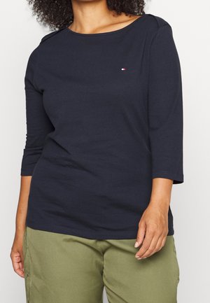 Femme portant un haut bleu marine à manches trois-quarts avec un petit logo sur la poitrine et un pantalon vert olive sur fond neutre.