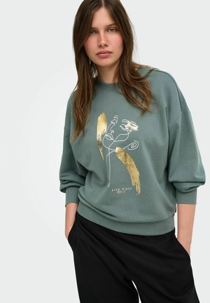 Jeune femme portant un sweat-shirt gris-vert avec un motif abstrait de visage doré et blanc et un pantalon noir, debout les mains dans les poches.