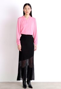Pink long-sleeve blouse with a V-neck, paired with a black pleated maxi skirt. Model wears black heeled boots. Plain background.